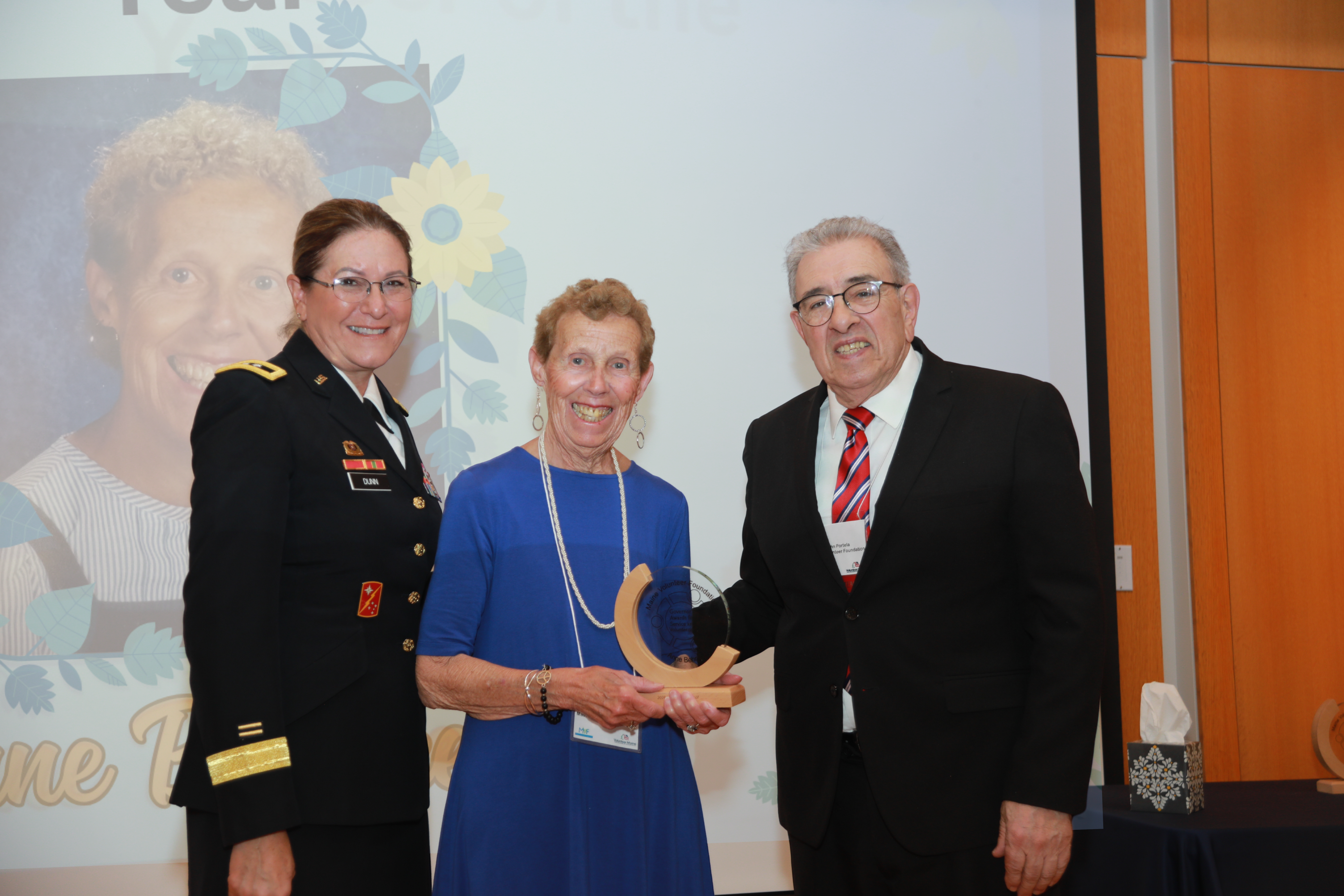 2026 Maine Volunteer of the Year award recipient Anne Behnke holding a crescent wood award standing in between Major General Diane Dunn and Maine Volunteer Foundation President John Portela
