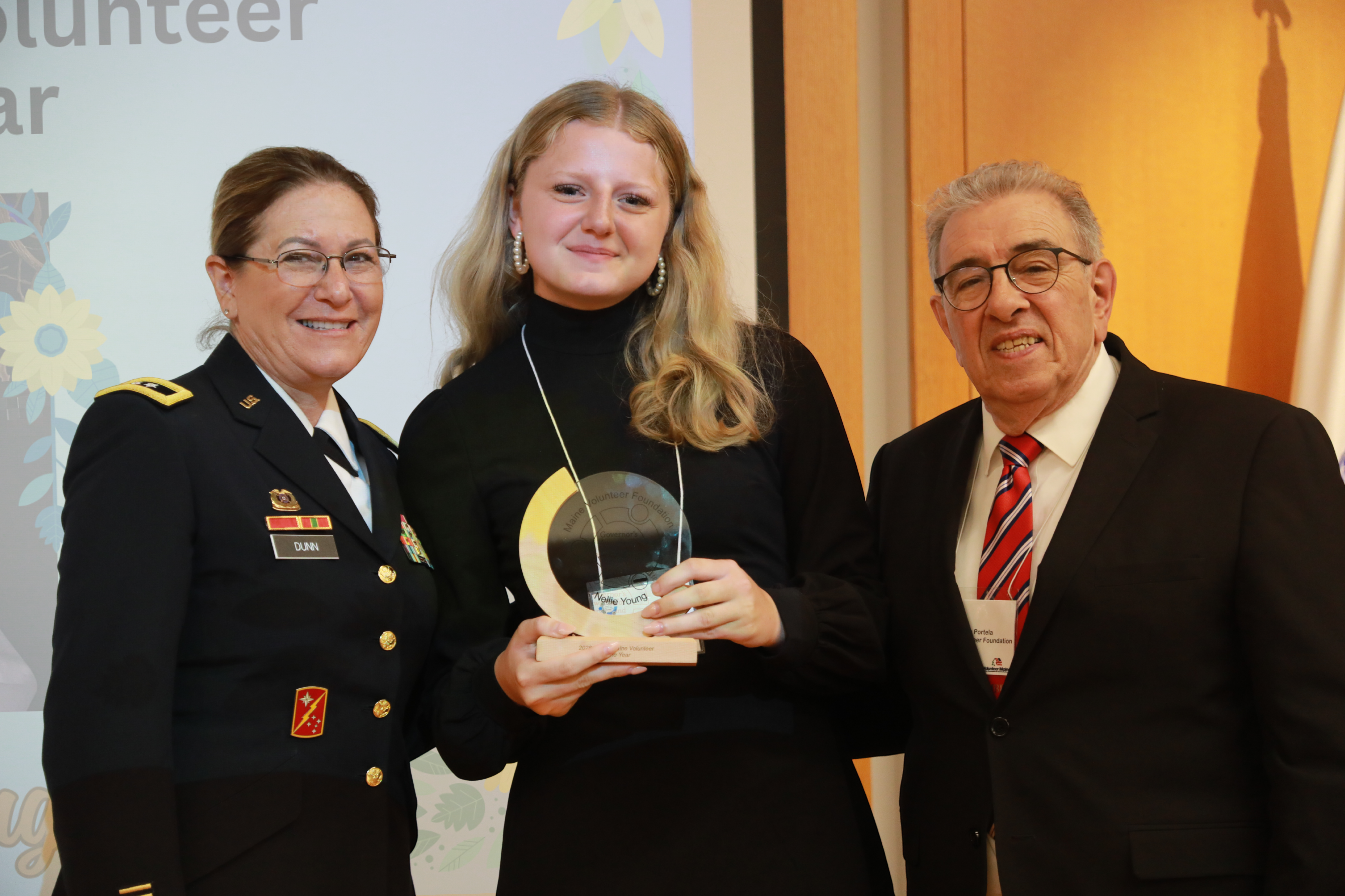 2026 Young Maine Volunteer of the Year awardee Nellie Young stands with a crescent wood and glass award in between Major General Diane Dunn and Maine Volunteer Foundation President John Portela