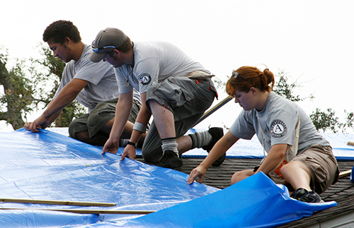 three AmeriCorps members up on a roof spreading a blue tarp on damaged room